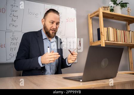 Männlicher Lehrer, der Englisch unterrichtet, auf einem Whiteboard mit grundlegenden Grammatikregeln sitzt, den Schülern neues Material erklärt. Online-Fremdsprachen Stockfoto