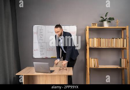 Konzept der Fernschulung. Glücklicher männlicher Lehrer, der im Home Office online Englisch Unterricht mit Laptop und Tafel mit Grammatikregeln gibt. Stockfoto