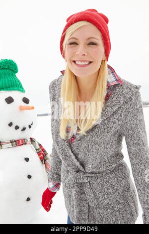 Ihr Lächeln erwärmt das Winterwunderland. Eine wunderschöne junge Frau, die neben einem Schneemann steht. Stockfoto