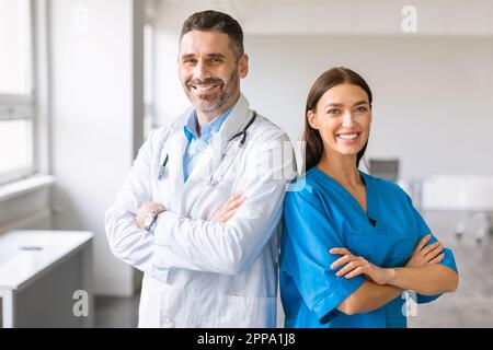 Selbstbewusste weiße Kollegen, männlicher Arzt und Krankenschwester, trägt Mäntel, posiert mit gefalteten Armen, lächelt vor die Kamera Stockfoto