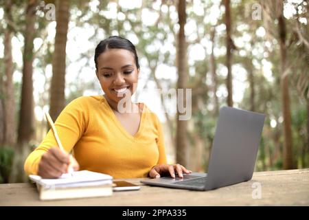 Glückliche brasilianische Studentin in ungezwungener Atmosphäre, die ihren Laptop benutzt und Notizen macht, ferngesteuert im Park lernt, draußen Stockfoto