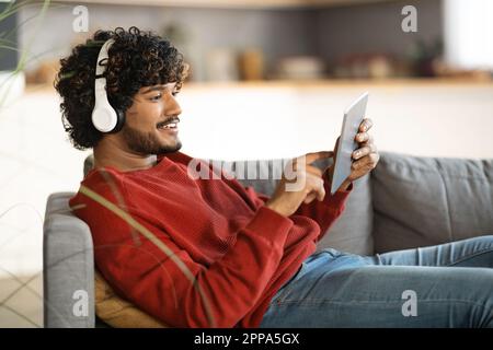 Lächelnder Indischer Mann Mit Kabellosen Kopfhörern Und Digitalem Tablet Zu Hause Stockfoto