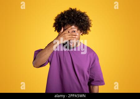 Trauriger schockierter erwachsener afroamerikanischer Lockenkopf in lila T-Shirt bedeckt Gesicht mit Händen Stockfoto