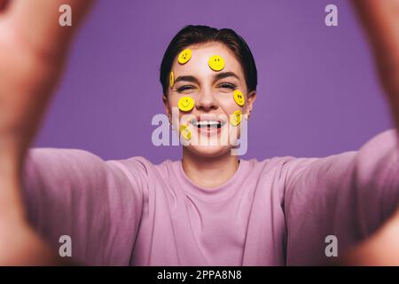 Verspielte junge Frau, die ein Selfie mit lächelnden Aufklebern im Gesicht macht. Glückliche Frau, die in die Kamera lächelt, während sie ein Foto von sich selbst macht. Fröhlicher Yo Stockfoto