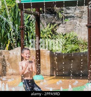 Asiatischer kleiner Junge, der im Sommer mit dem Wasser im Brunnen des Parks spielt Stockfoto