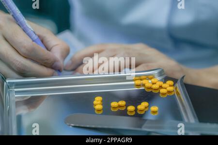 Gelbe zuckerbeschichtete Tablette auf Edelstahltablett. Apothekerhandhalter und das Schreiben von Arzneimittelanweisungen auf Plastikpillen-Packungen. Prescrip Stockfoto