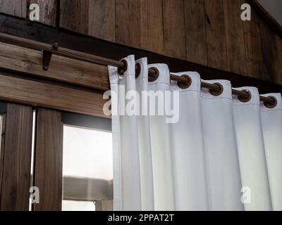 Nahaufnahme eines weißen Wandvorhangs an einer Holzvorhangschiene, die an der Holzplankenwand des tropischen Giebelgebäudes in der Nähe der Glasschiebetür dekoriert ist Stockfoto