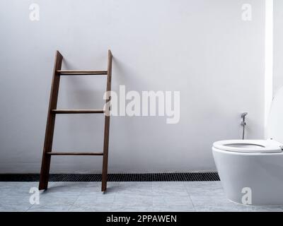 Weiße Toiletteneinrichtung, minimalistischer Stil. Handtuchhalter aus Holz, dunkelbraune Stehleiter für Handtücher in der Nähe der Toilette mit Keramikspülung und Bidet-Dusche auf dem Platz Stockfoto