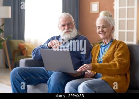 Glückliches Seniorenpaar, das sich etwas auf dem Laptop ansieht und lächelt, während es auf dem Sofa im Wohnzimmer sitzt Stockfoto
