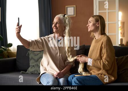 Glückliche Seniormutter, die zusammen mit ihrer erwachsenen Tochter Selfie-Porträt auf ihrem Smartphone macht, während sie ihr auf dem Sofa eine Frisur macht Stockfoto