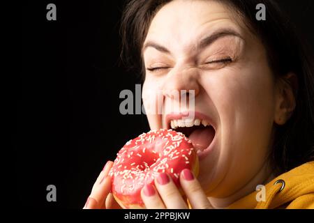 Eine Frau beißt einen großen roten Donut, einen schwarzen Hintergrund, ein Ort für Text. Gluttonie, überfressen und zuckersüchtig. Stockfoto