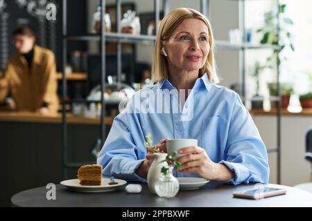 Glückliche blonde, reife Frau mit Kopfhörern, die Musik hört, während sie im Café am Tisch sitzt und eine Tasse Tee oder Kaffee mit Kuchen trinkt Stockfoto
