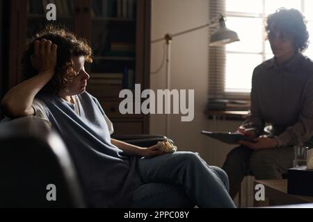 Traurige junge brünette Patientin einer Beraterin mit Gewebe in der Hand, die auf der Couch sitzt und ihr Problem dem Psychotherapeuten in der Praxis beschreibt Stockfoto