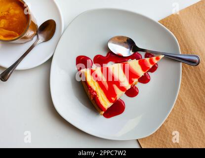 Ein Stück Käsekuchen mit frischer Erdbeermarmelade, serviert auf einem grauen Teller neben einem Glas Kaffee in der Cafeteria Stockfoto