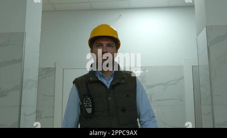 Nahaufnahme des weißen männlichen Konstrukteurs in gelbem Helm geht im Gebäude und macht Walkie-Talkie auf der Baustelle. Gut aussehender Mann Stockfoto