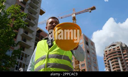 Blick von unten auf einen freudigen jungen Bauarbeiter, der vor der Baustelle steht und nach draußen blickt. Schließen. Männlicher Ingenieur in Uniform setzt Helm auf Stockfoto