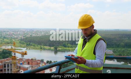 Fokussierter junger weißer Mann in Uniform und Helm, der auf einer Baustelle auf einem digitalen Tablet arbeitet. Kompetenter Ingenieur, der die Arbeit überprüft Stockfoto