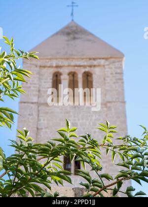 Rab, Kroatien - 24. August 2022: Ein atemberaubender Glockenturm einer Kirche in Rab, Kroatien, sticht vor dem blauen Himmel hervor. Blattbäume und Pflanzen säumen die Mauer Stockfoto