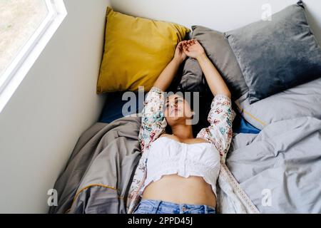Nachdenkliche Frau mit erhobenen Armen, die zu Hause auf dem Bett lag Stockfoto