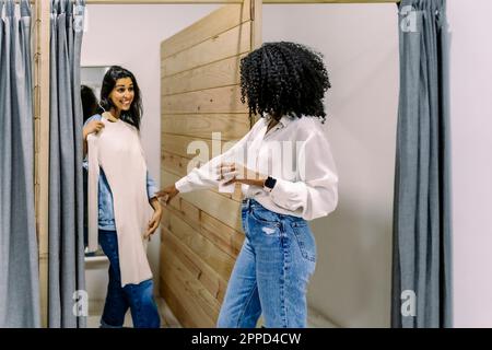 Glückliche junge Frau mit einer Freundin, die Kleidung in der Umkleidekabine probiert Stockfoto