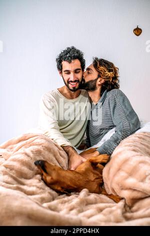Schwuler Mann, der seinen Freund küsst und zu Hause mit dem Hund im Bett spielt Stockfoto