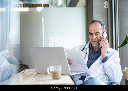 Arzt prüft medizinischen Bericht und diskutiert auf Smartphone am Schreibtisch Stockfoto