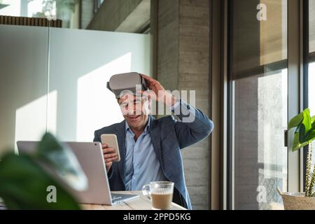 Ein erwachsener Geschäftsmann mit VR-Brille, der im Büro ein Smartphone benutzt Stockfoto