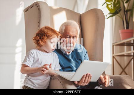 Großvater, der zu Hause in einem Sessel sitzt und seinen Laptop teilt Stockfoto