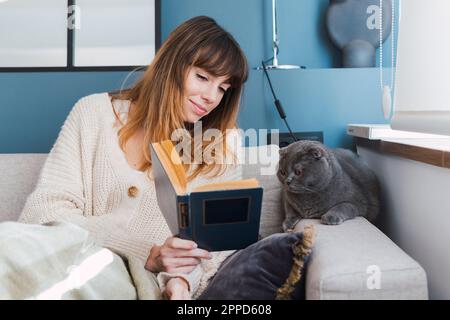 Frau, die der Katze ein Buch zeigt, sitzt zu Hause auf dem Sofa Stockfoto