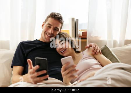 Glückliches Paar mit Smartphones, das sich zu Hause im Bett entspannen kann Stockfoto