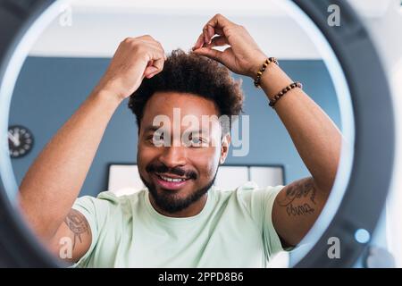 Lächelnder Mann mit Hand im Haar hinter Ringlicht zu Hause Stockfoto