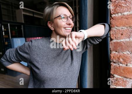 Glückliche reife Frau, die durch eine Smartwatch spricht, die sich an die Wand lehnt Stockfoto