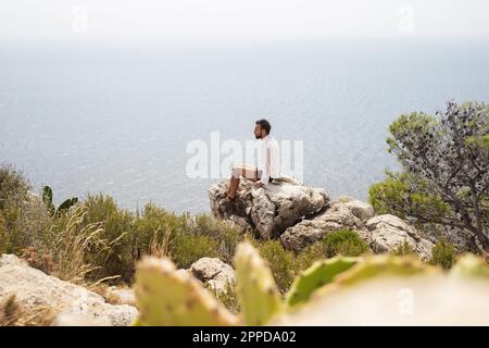 Junger Mann, der auf einem Felsen in der Nähe des Meeres sitzt Stockfoto