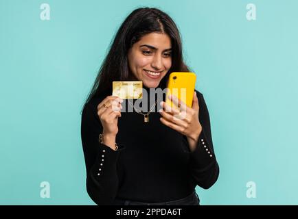 Ehrliche, indische Kundin, die bei Geldüberweisungen, Einkäufen im Internet und Bezahlung eine Kreditkarte und ein Smartphone verwendet. Finanzen und Internet. Hübsches Mädchen, isoliert auf blauem Hintergrund Stockfoto