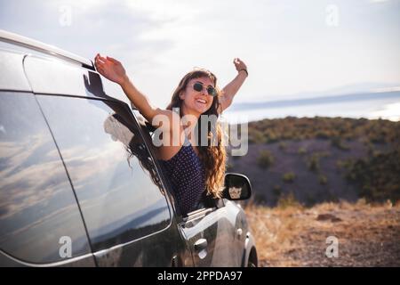 Glückliche junge Frau mit erhobenen Armen, die sich aus dem Autofenster lehnen Stockfoto