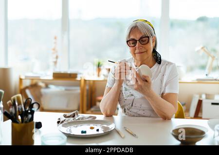 Lächelnde Seniorin malt Becher im Studio Stockfoto