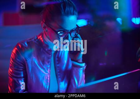 Neon-Metaverse-futuristisches Konzept. Pensive, elegante 40-jährige Frau mit Brille und Laptop, die in einem modernen Büro über ein Smartphone spricht. Stockfoto