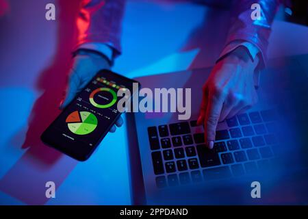 Neon-Metaverse-futuristisches Konzept. Nahaufnahme einer Frau mit Laptop und Smartphone, die im Büro Grafiken untersucht. Stockfoto