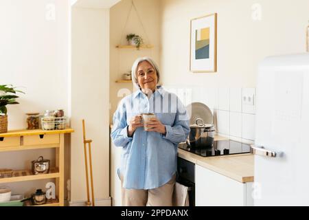 Lächelnde Seniorin, die mit einem Becher in der Küche steht Stockfoto
