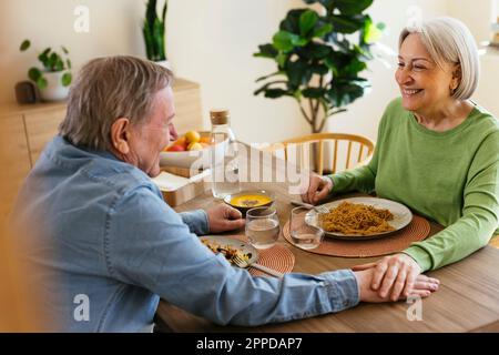 Glückliches Seniorenpaar mit gestapelten Händen, das zu Hause zu Mittag isst Stockfoto