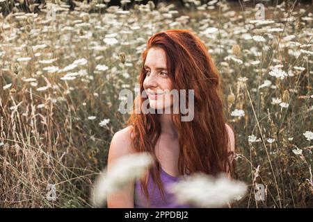 Lächelnde rothaarige junge Frau inmitten weißer Blumen auf dem Feld Stockfoto