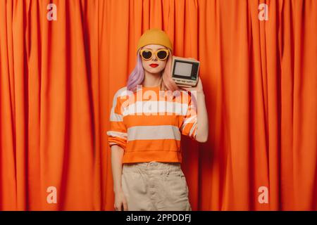 Frau mit Strickmütze, die einen Fernseher im Retro-Stil vor dem orangefarbenen Vorhang hält Stockfoto