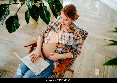 Schwangere Frau, die einen Laptop benutzt und zu Hause auf einem Stuhl sitzt Stockfoto