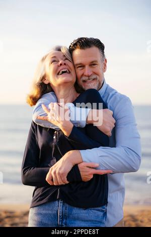Ein glücklicher Mann, der eine fröhliche, reife Frau am Strand umarmt Stockfoto