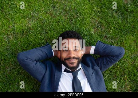 Rücksichtsvoller lächelnder Geschäftsmann mit Händen hinter dem Kopf, der auf dem Gras liegt Stockfoto