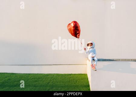 Ein Mädchen als Astronaut verkleidet, sitzt an der Wand und hält einen herzförmigen Ballon Stockfoto