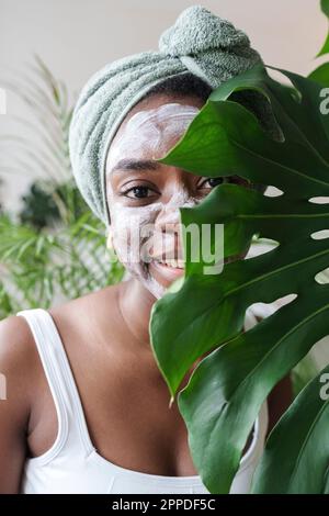 Lächelnde Frau mit Gesichtsmaske hinter Monstera-Blatt zu Hause Stockfoto