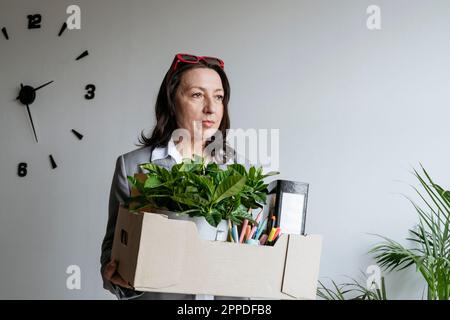 Traurige Geschäftsfrau, die eine Kiste mit Büromaterial vor der Wand hält Stockfoto