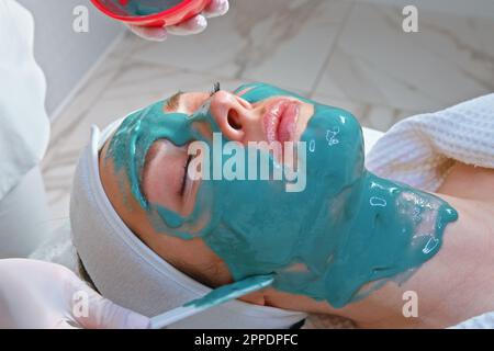 Eine Kosmetikerin, die eine Maske auf das Gesicht einer jungen liegenden Frau in einem Schönheitssalon aufträgt, um eine Hautreinigung und Feuchtigkeitsbehandlung durchzuführen. Stockfoto