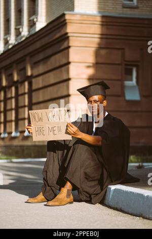 Ein glücklicher Schwarzer sitzt mit einem Plakat auf der Straße und sucht einen Job. Universitäts- oder Universitätsstudenten mit Graduiertenkleid und -Mütze. Studieren Sie und Stockfoto